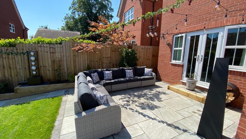 Grey porcelain patio with a rattan corner sofa, festoon lights and a lawn edged in sleepers