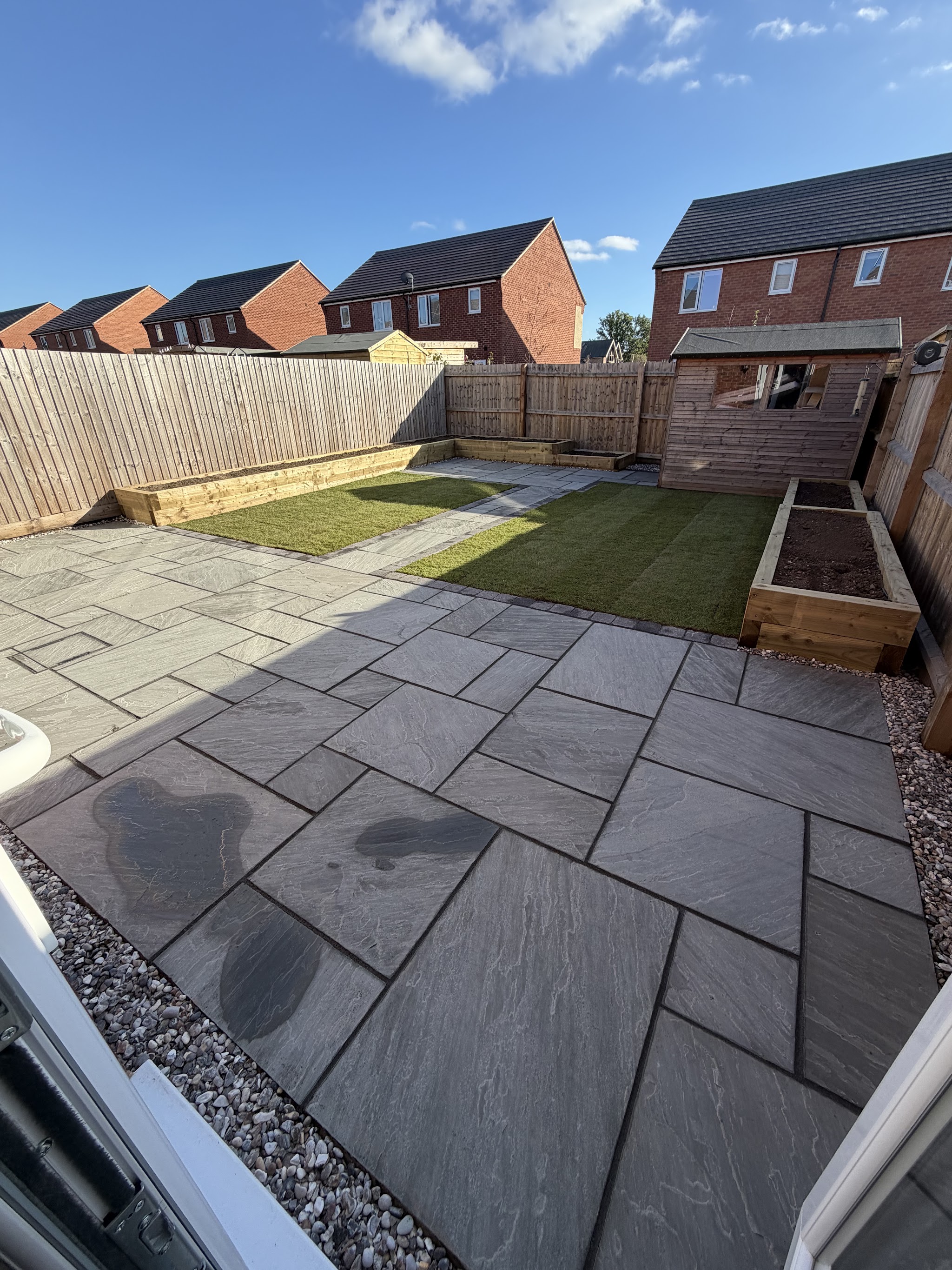 Dark grey patio with a gravel border, lawn and sleeper raised beds behind a new-build house